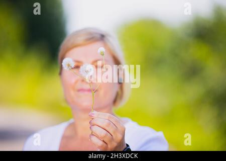 Verschwommenes Foto einer blonden Frau im Freien, die die Nelkenglüte in der Hand hält und eine smarte Uhr und Kopfhörer trägt. Stockfoto