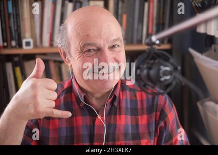 Älterer pensionierter Mann blogger, der sich die Kamera ansieht, die Vlog aufzeichnet, und eine „Call Me“-Geste zeigt. Stockfoto