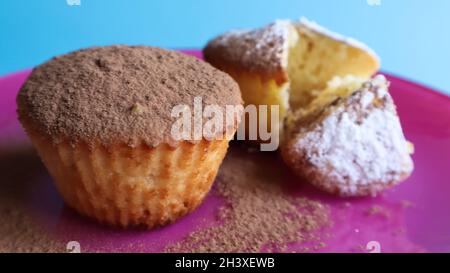 Zwei mit Schokolade und Puderzucker bestreute Quark-Kuchen auf einem rosa Teller auf blauem Hintergrund. Dessert, ein kleiner Cupcake. Weiß b Stockfoto
