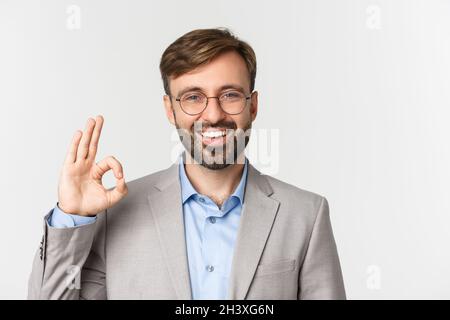 Nahaufnahme eines gutaussehenden bärtigen Mannes in grauem Anzug und Brille, etwas empfehlen, Zeichen in Ordnung zeigen und lächeln, Zustimmung geben, st Stockfoto