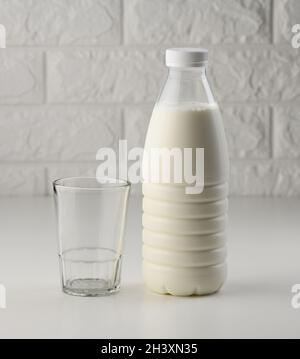 Volle Plastikmilchflasche und leere Glasbecher auf weißem Tisch, Backsteinwand Hintergrund Stockfoto