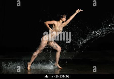 Junge schöne Frau mit kaukasischem Aussehen und langen Haaren tanzt in Wassertropfen auf schwarzem Hintergrund. Springen und Schwingen Stockfoto