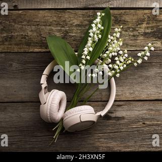 Beigefarbene kabellose Kopfhörer auf einem Holztisch, neben einem Bouquet aus weißen Lilien des Tals Stockfoto