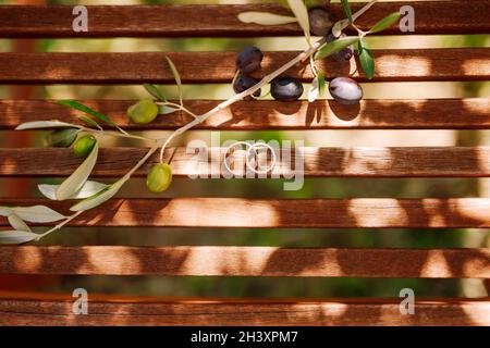 Draufsicht auf goldene Eheringe auf Holzplanken mit einem Zweig Olivenbaum mit Früchten im Sonnenlicht. Stockfoto