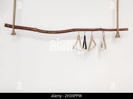 Fünf leere Kleiderbügel, die an einem dicken Seil, isoliert über einer weißen Wand, an einem rustikalen Holzstab hängen Stockfoto