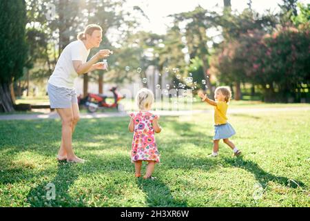 Frau bläst Seifenblasen, und zwei kleine Mädchen fangen sie auf einer Lichtung Stockfoto