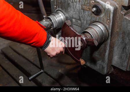 Die Hand eines Mannes in einem roten Arbeitsanzug hält sich am Griff fest und steuert die Arbeit einer alten Fräsmaschine in einer Industrieanlage. Stockfoto