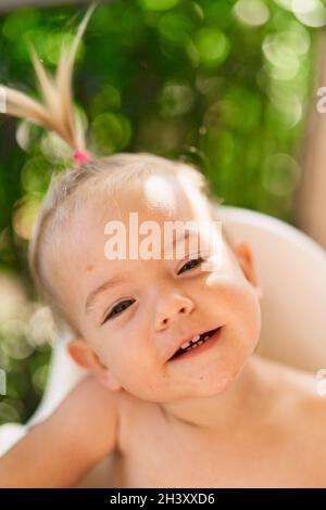 Lächelndes kleines Mädchen mit Pferdeschwanz sitzt auf einem Hochstuhl. Hochformat Stockfoto