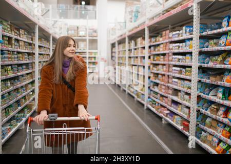 Junge Frau mit Lebensmittelkarren und Regale mit Lebensmitteln in einem Geschäft. Stockfoto