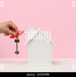 Hand hält Schlüssel und Holzhaus auf einem rosa Hintergrund. Immobilien Vermietung, Kauf und Verkauf Konzept Stockfoto