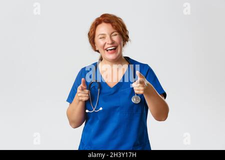 Medizin, Gesundheitswesen und Coronavirus-Konzept. Fröhliche, enthusiastische rothaarige Ärztin, Ärztin in Scrubs, die auf Fi zeigen Stockfoto
