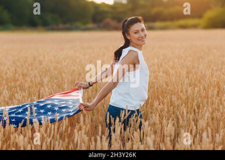 Frau mit amerikanischer Flagge im Weizenfeld bei Sonnenuntergang. Juli. Unabhängigkeitstag patriotischer Feiertag. Stockfoto