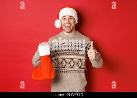 Konzept der Winterferien, Neujahr und Feier. Fröhlicher, gutaussehender Mann mit Weihnachtsmütze und Pullover, der Weihnachtsstrumpf zeigt Stockfoto