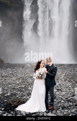 Hochzeitspaar in der Nähe des Skogafoss Wasserfalls. Reiseziel Island Hochzeit. Der Bräutigam umarmt die Braut an den Schultern, sie lachen zusammen Stockfoto