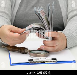 Ein Stapel von hundert Dollar Scheinen in der Hand einer Frau und eine hölzerne Lupe auf einem weißen Tisch. Budgetanalyse, Einkommen und Stockfoto