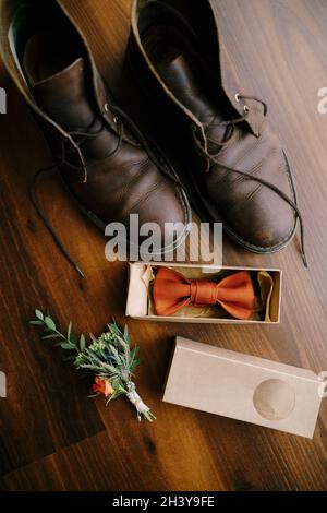 Fliege in einer Box für einen Bräutigam mit Boutonniere und Herrenstiefel mit ungebundenen Schnürsenkeln auf braunem Boden. Stockfoto