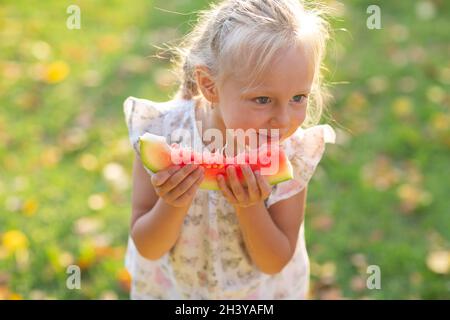 Cute blonde kleine Mädchen essen Wassermelone auf dem Gras im Park Stockfoto