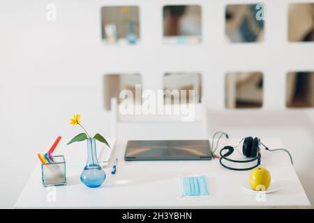 Moderner Innenarbeitsplatz mit silbernem Laptop, Kopfhörer, Vase, Blume, Apfel, Gesichtsmaske und Stifte auf dem Tisch. Stockfoto