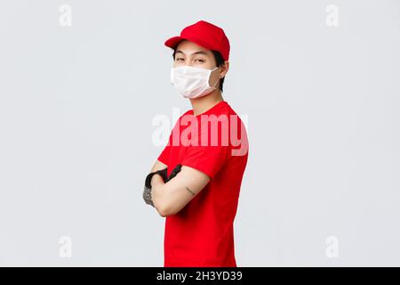 Selbstbewusster asiatischer Lieferer in roter Mütze, medizinischer Maske und Handschuhen, Standprofil mit gekreuzten Armen auf der Brust, Kamera mit drehen Stockfoto