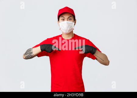 Schockiert und verhört asiatischen Delivery Mann in Uniform roten Kappe und T-Shirt, trägt medizinische Schutzmaske und Handschuhe. Liefern Sie Stockfoto