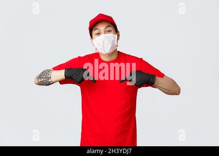 In der Zeit Lieferung mit dieser Firma Kuriere Service. Fröhlicher asiatischer Kurier in Mütze und rotem T-Shirt, mit medizinischer Maske und glo Stockfoto