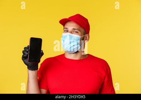 Covid-19, Lieferaufträge, Shopping, kontaktloses Bezahlen und soziales Distanzierungskonzept. Schöner Kurier in roter Uniform und medi Stockfoto