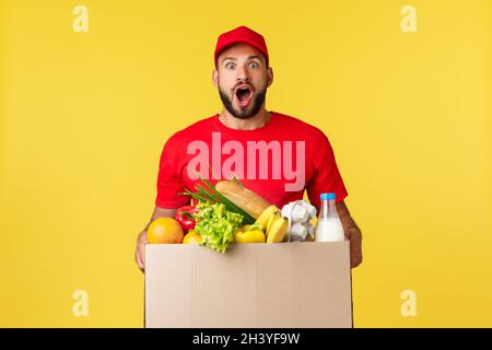 Online-Lieferung, Shopping und Food Order Konzept. Aufgeregt und amüsiert Kurier in roter Uniform, bärtige Lieferung Mann keuchte astonis Stockfoto