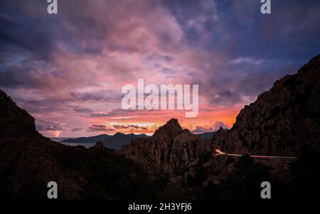 Sonnenaufgang über den Calanches von Piana auf Korsika mit Blitz in der Ferne und Autoampeln, die entlang der D81 fahren Stockfoto