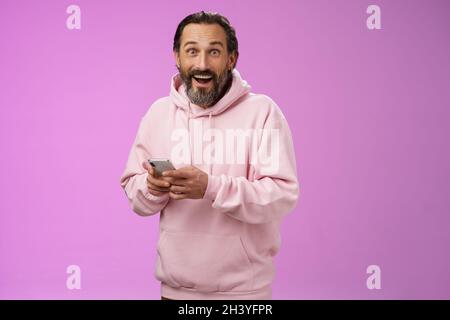 Amüsiert glücklich fasziniert erwachsenen reifen kaukasischen bärtigen Mann in rosa Hoodie keuchend beeindruckt halten Smartphone aussehen Kamera aston Stockfoto