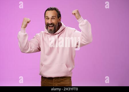 Ja, wer kümmert sich um das Alter. Sorglos glücklich glücklich alten Mann bärtig in trendigen rosa Kapuzenpullover heben Fäuste freudig triumphieren mit Spaß gl Stockfoto