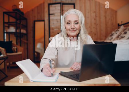Ältere Frau mit Kopfhörern, die in der Küche Videoanruf auf dem Laptop spricht, am Bildschirm winkt, chattet und das Fotogerät spricht Stockfoto