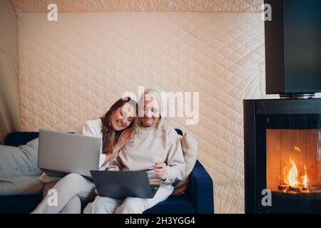 Ältere und junge Frau mit Computer-Laptop auf dem Sofa. Mutter und Tochter sitzen drinnen am Kamin Stockfoto