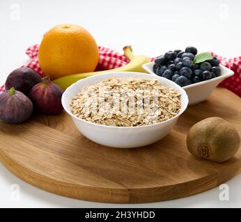 Rohe Haferflocken in weißen Keramikplatte und Obst auf weißem Tisch, Frühstück Stockfoto