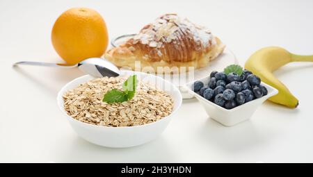 Rohe Haferflocken in weißer Keramikplatte, Heidelbeeren, Orange, Banane, auf weißem Tisch, Frühstück Stockfoto