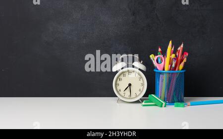 Weißer runder Wecker und ein Metallglas mit Stiften, Bleistiften und Filzstiften auf dem Hintergrund einer leeren schwarzen Kreidetafel Stockfoto