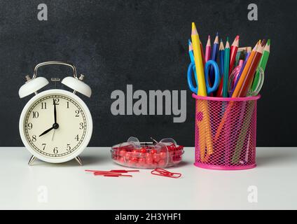 Weißer runder Wecker und ein Metallglas mit Stiften, Bleistiften und Filzstiften auf dem Hintergrund einer leeren schwarzen Kreidetafel Stockfoto