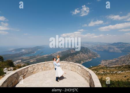 Der Bräutigam hält die Braut auf der Aussichtsplattform in den Armen, dahinter ein Panoramablick auf die Bucht von Kotor Stockfoto