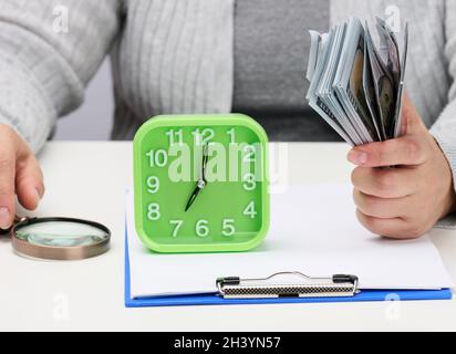 Stapel von hundert Dollar Scheinen in der Hand einer Frau und eine hölzerne Lupe auf einem weißen Tisch. Budgetanalyse, Einkommen und Stockfoto