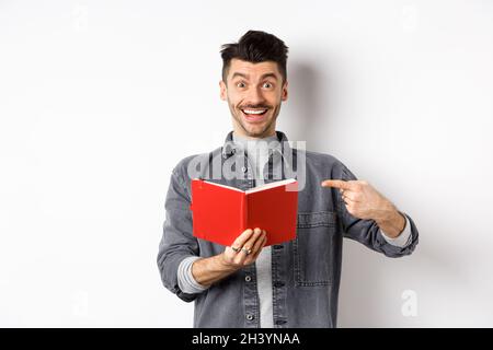 Glücklicher lächelnder Kerl zeigt auf roten Planer, zeigt gute Zeitschrift oder Buch, auf weißem Hintergrund stehen Stockfoto