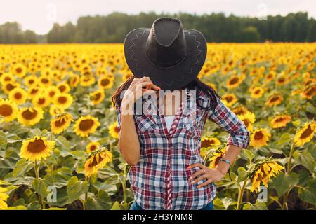 Junge Frau Agronomin trägt Cowboyhut, kariertes Hemd und Jeans auf Sonnenblumenfeld. Umfassendes Erntekonzept. Stockfoto