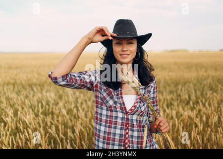 Die amerikanische Farmerin lächelt mit den Weizenohren in Cowboyhut, karierten Hemden und Jeans auf dem Weizenfeld Stockfoto