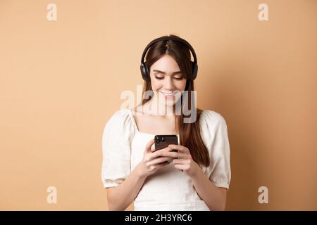 Lächelnde Frau, die Nachrichten am Telefon schreibt und Musik über kabellose Kopfhörer hört, Videos auf dem Smartphone ansieht, beige Hintergrundmusik Stockfoto