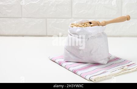 Roher Haferbrei in einem weißen Papierbeutel und ein Holzlöffel auf einem weißen Tisch, Frühstücksbrei Stockfoto