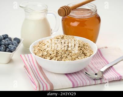 Frühstück am Morgen, rohe Haferflocken auf einem Keramikplatte, Milch in einem Dekanter und Honig in einem Glas auf einem weißen Tisch Stockfoto