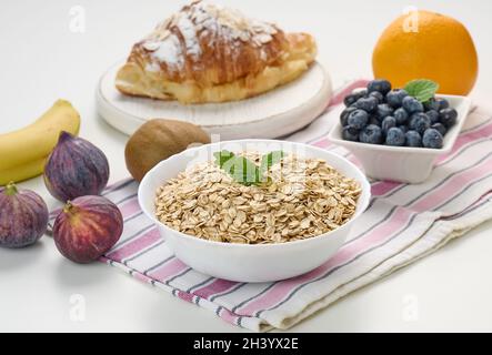 Rohe Haferflocken in weißer Keramikplatte, Heidelbeeren, Feigen, Orange, Banane, Auf weißem Tisch Frühstück Stockfoto