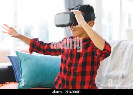 Lächelnder asiatischer Junge, der auf der Couch sitzt und ein vr-Headset benutzt Stockfoto