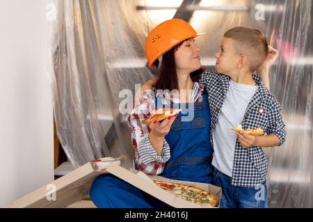 Entzückende Familie Mutter und Sohn ruhen sich aus und essen leckere italienische Pizza müde nach der Renovierung in einem neuen Haus. Kaukasische Frau in Schutzhelm Stockfoto