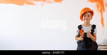 Ein kleiner Junge mit einem Pinsel auf dem Hintergrund einer weißen Wand mit einem orangefarbenen Fleck zeigt die Klasse. Im Zimmer werden gerade Renovierungsarbeiten für Kinder durchgeführt. Stockfoto