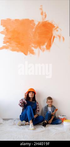 Renovierung in der Wohnung. Glückliche Familie Mutter und Sohn bemalten die Wand mit oranger Farbe. Mama im Helm und der Junge auf dem Holzboden sitzen Stockfoto