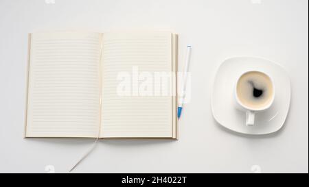 Offenes Notizbuch mit leeren weißen Laken und einer Tasse Kaffee auf einem weißen Tisch, Arbeitsplatz, Draufsicht Stockfoto